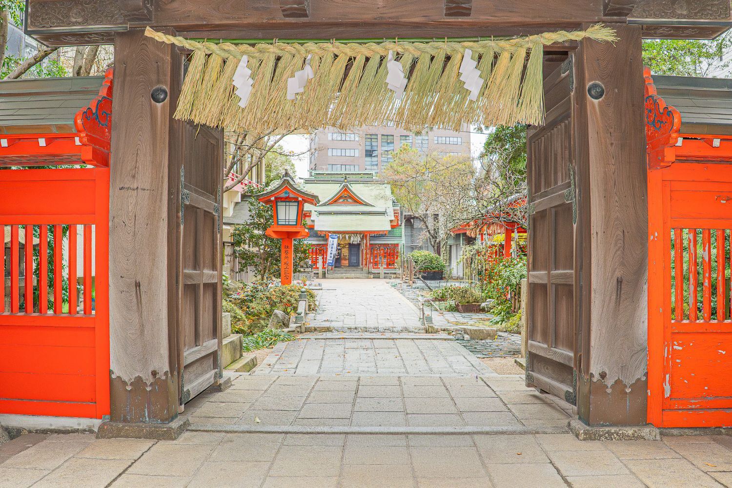 Wooden shrine gate with a straw shimenawa rope opening onto a vermilion shrine in Fukuoka
